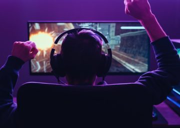 A gamer wearing headphones raises both fists while playing a video game on a monitor, with a glowing PC setup.