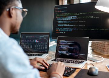 A man in a workplace wearing glasses and sitting in front of three screens. He uses a keyboard to write some code.