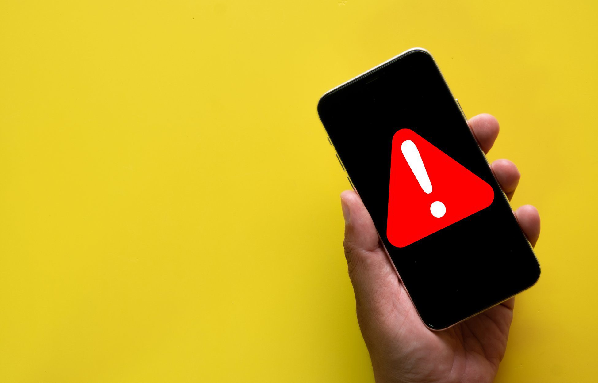 Against a yellow background, a hand holds up a smartphone that displays a warning exclamation point inside a red triangle.