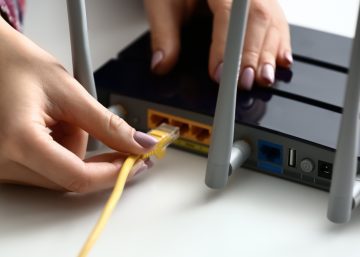 A close-up of a person plugging an Ethernet cable into a router box. The cable is yellow and the box is black.