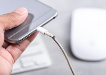 Someone holds a smartphone in their hand connected to its charger. The charging cable is frayed and broken at the connector.