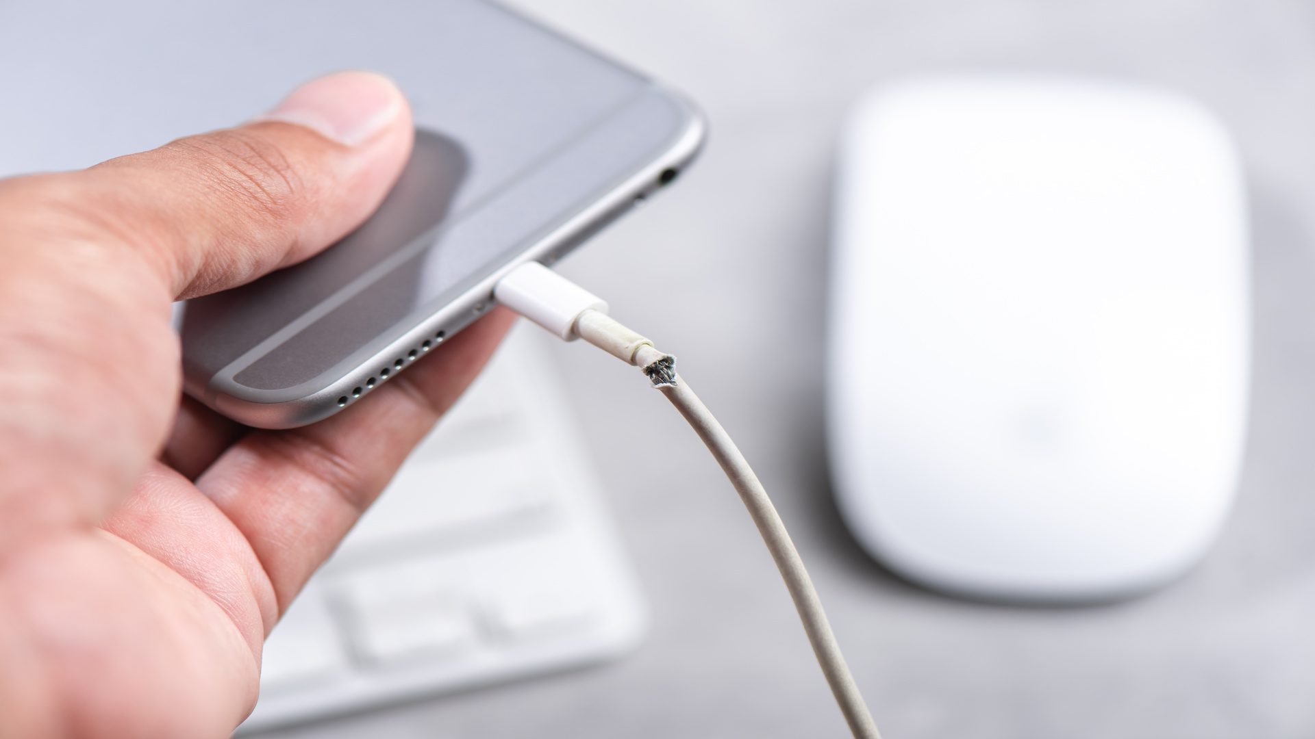 Someone holds a smartphone in their hand connected to its charger. The charging cable is frayed and broken at the connector.
