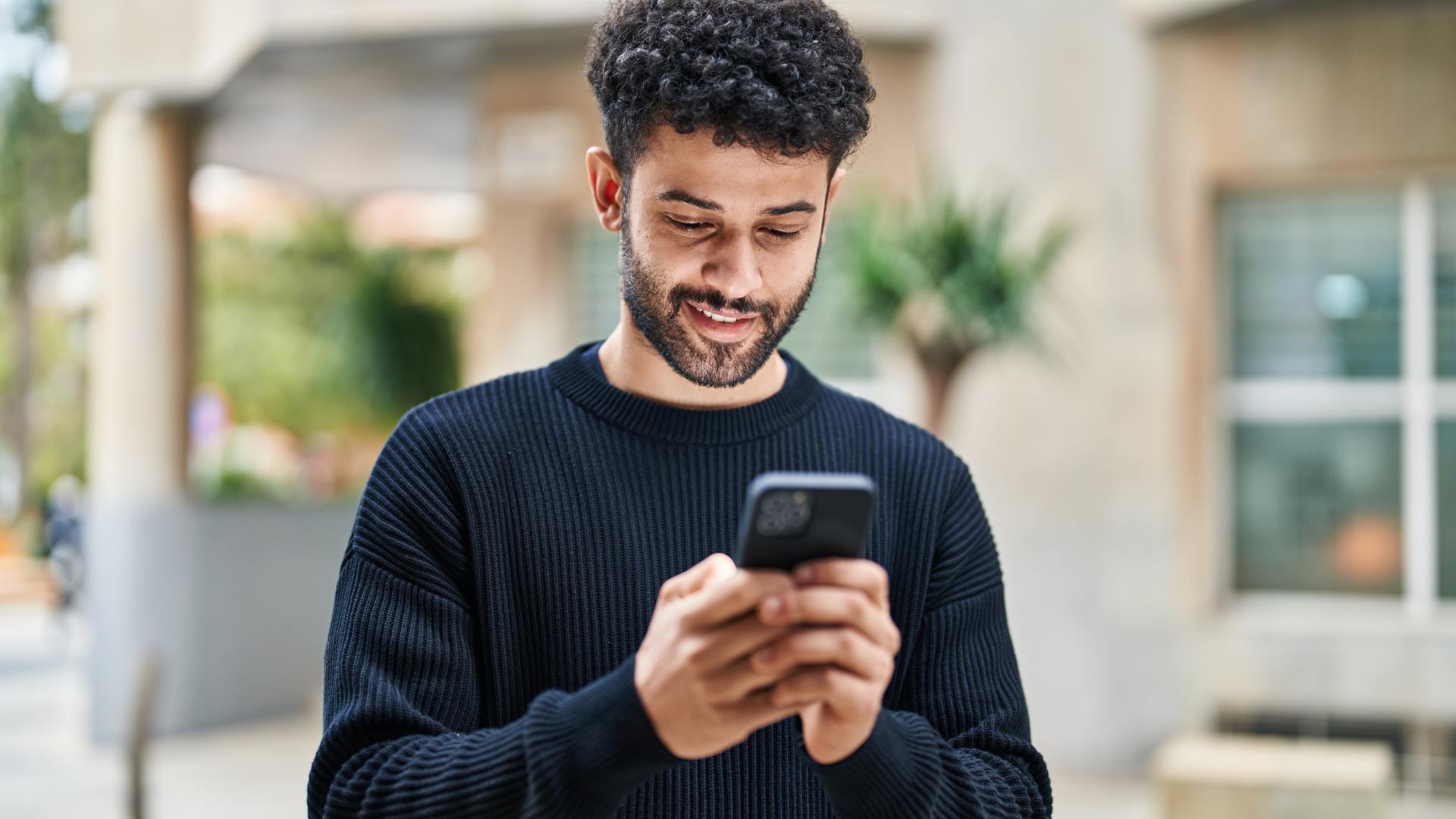 A younger, middle-aged man standing outside a house with his smartphone in both hands. He stares at the screen.