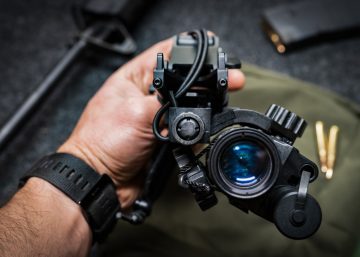 A hand is holding monocular night vision equipment with a helmet mount. A green bag is lying on a table in the background.