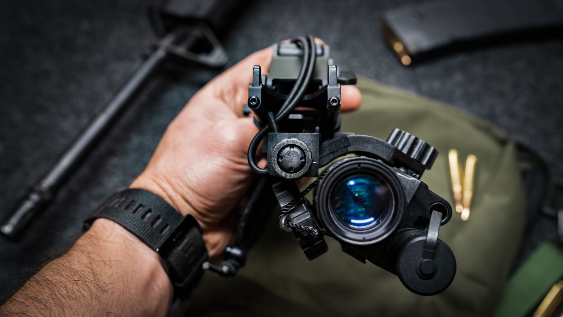 A hand is holding monocular night vision equipment with a helmet mount. A green bag is lying on a table in the background.