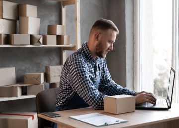 A man sits at a desk next to a window, typing on a laptop. A small cardboard box sits on the desk next to the laptop.
