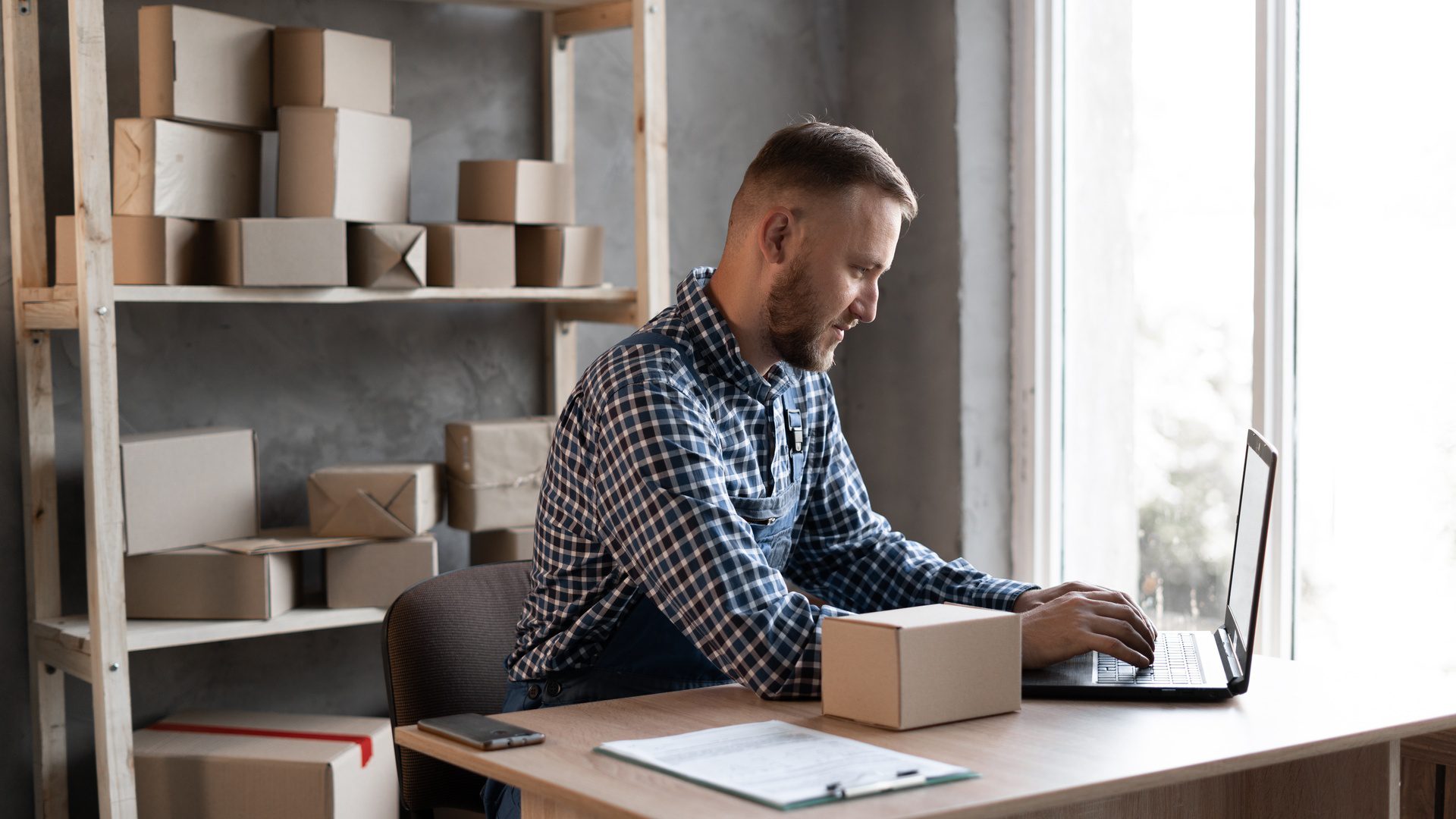 A man sits at a desk next to a window, typing on a laptop. A small cardboard box sits on the desk next to the laptop.