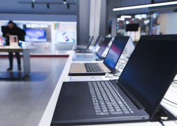 A row of open laptops on display in an electronics store. A customer shops at another display in the background.