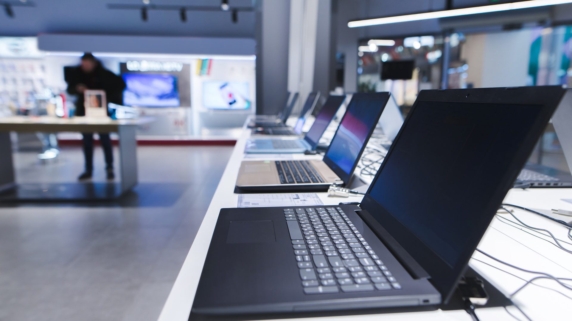 A row of open laptops on display in an electronics store. A customer shops at another display in the background.