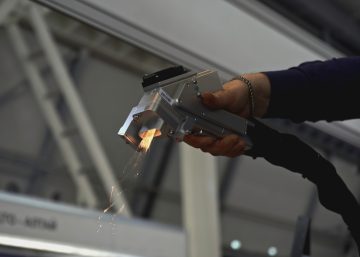 A person holding a handheld laser tool emitting sparks toward metal parts inside a large industrial interior.