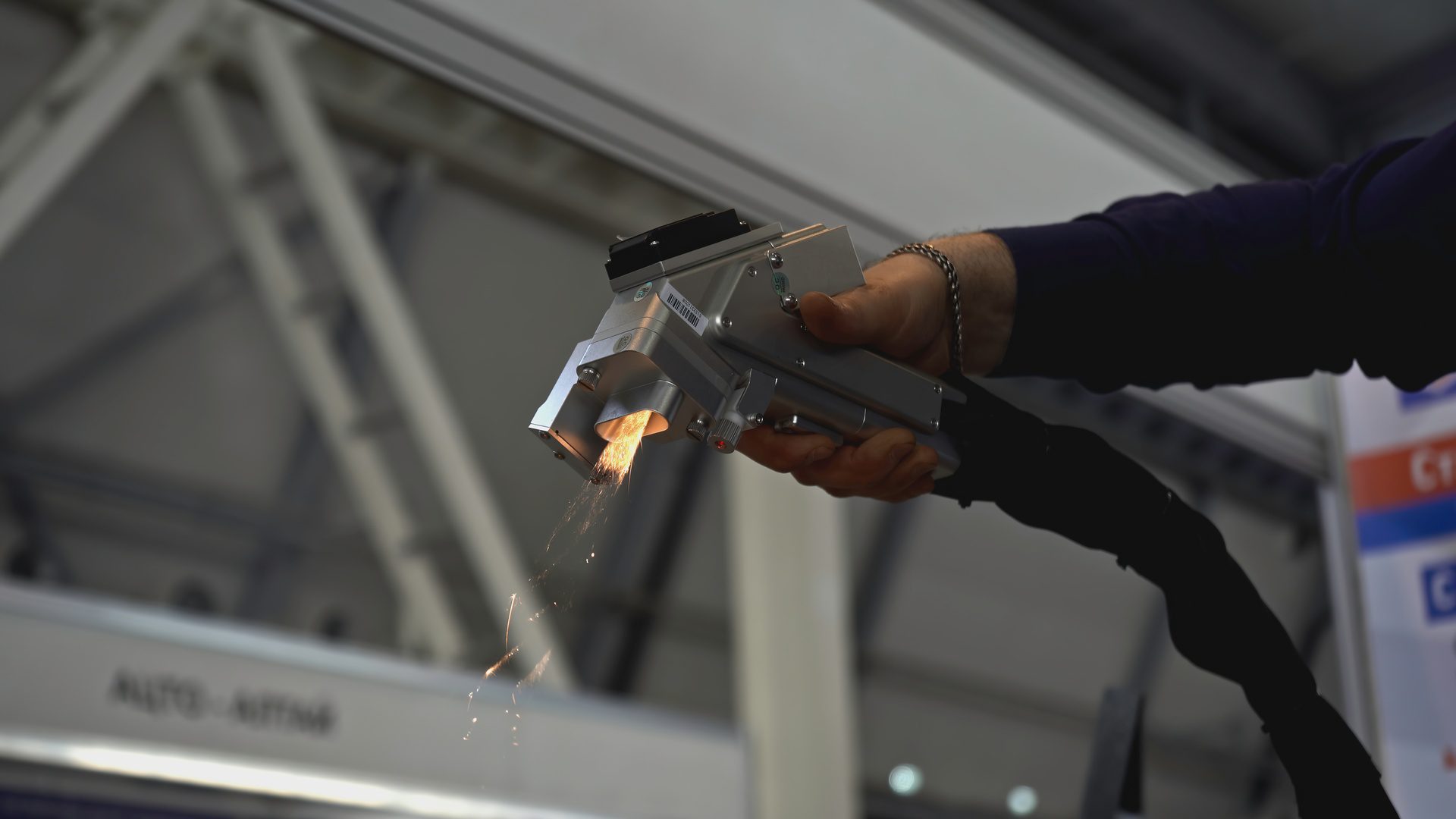 A person holding a handheld laser tool emitting sparks toward metal parts inside a large industrial interior.