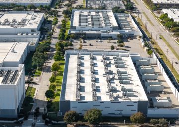 An aerial of white industrial buildings with rooftop equipment, tree-lined streets, and a few parked vehicles.