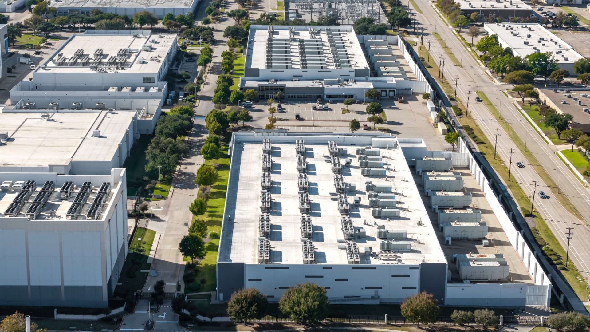 An aerial of white industrial buildings with rooftop equipment, tree-lined streets, and a few parked vehicles.