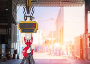 A digital crane scale hangs from a yellow hook above black lifting slings inside a sunlit industrial warehouse.