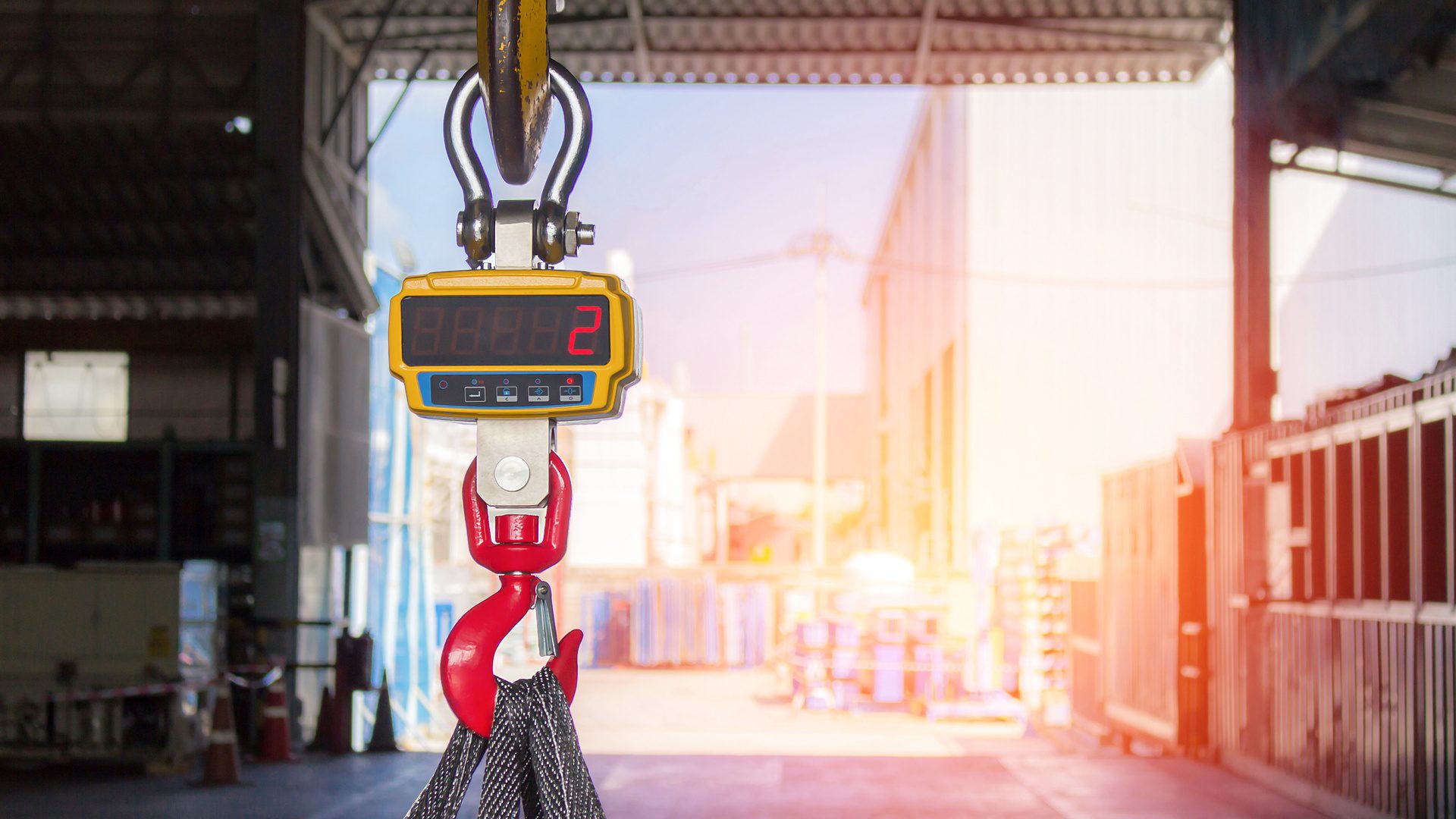 A digital crane scale hangs from a yellow hook above black lifting slings inside a sunlit industrial warehouse.