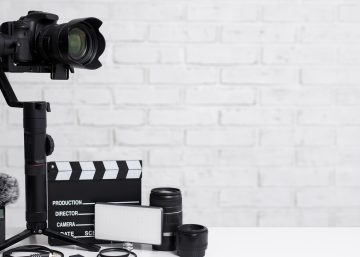 A camera is mounted on a stabilizer. A clapperboard, microphone, and camera lenses on lying on a white table.
