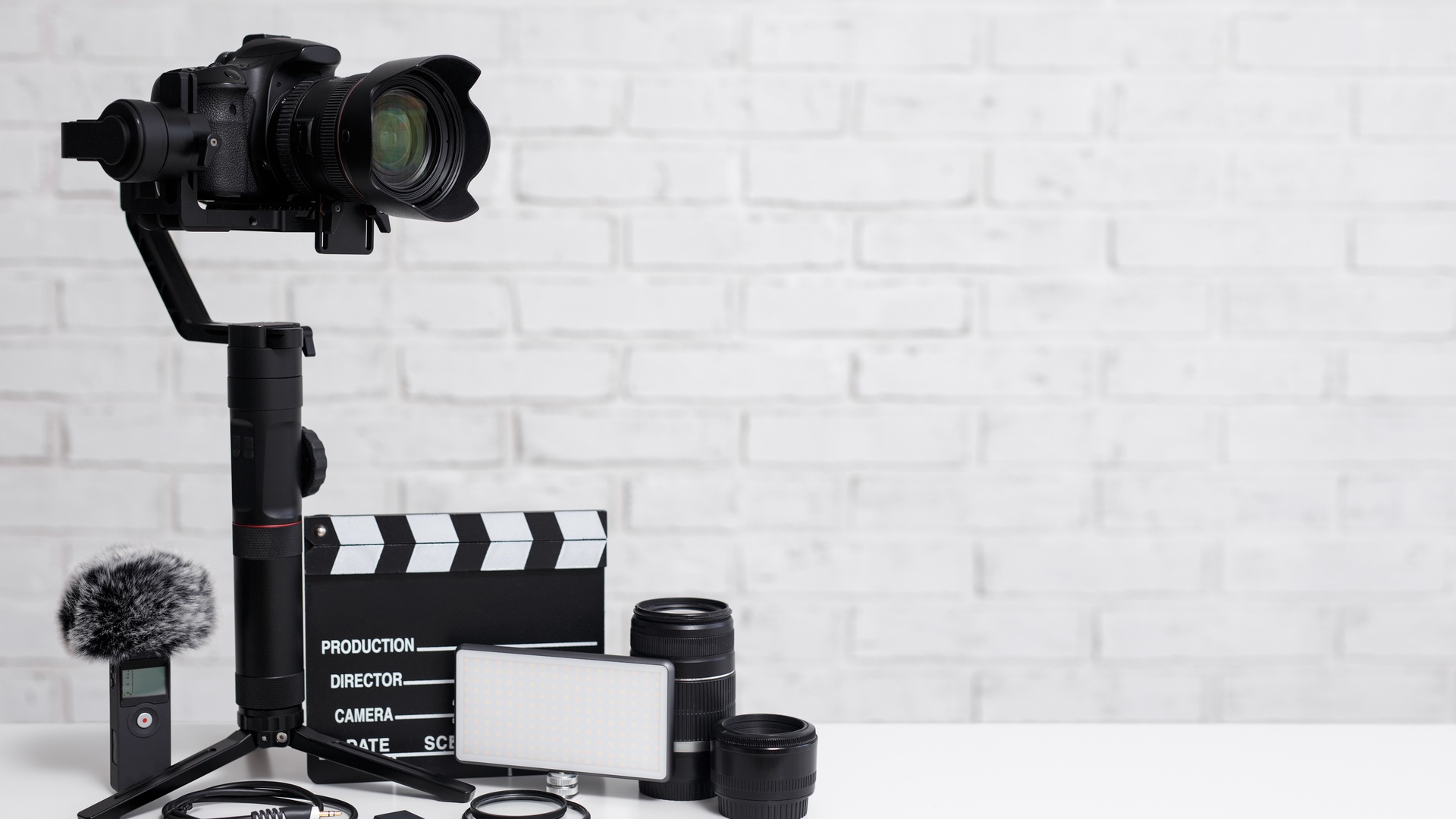 A camera is mounted on a stabilizer. A clapperboard, microphone, and camera lenses on lying on a white table.