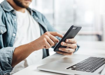 A person sits at a desk holding a black smartphone and scrolling. An open laptop sits in front of them.