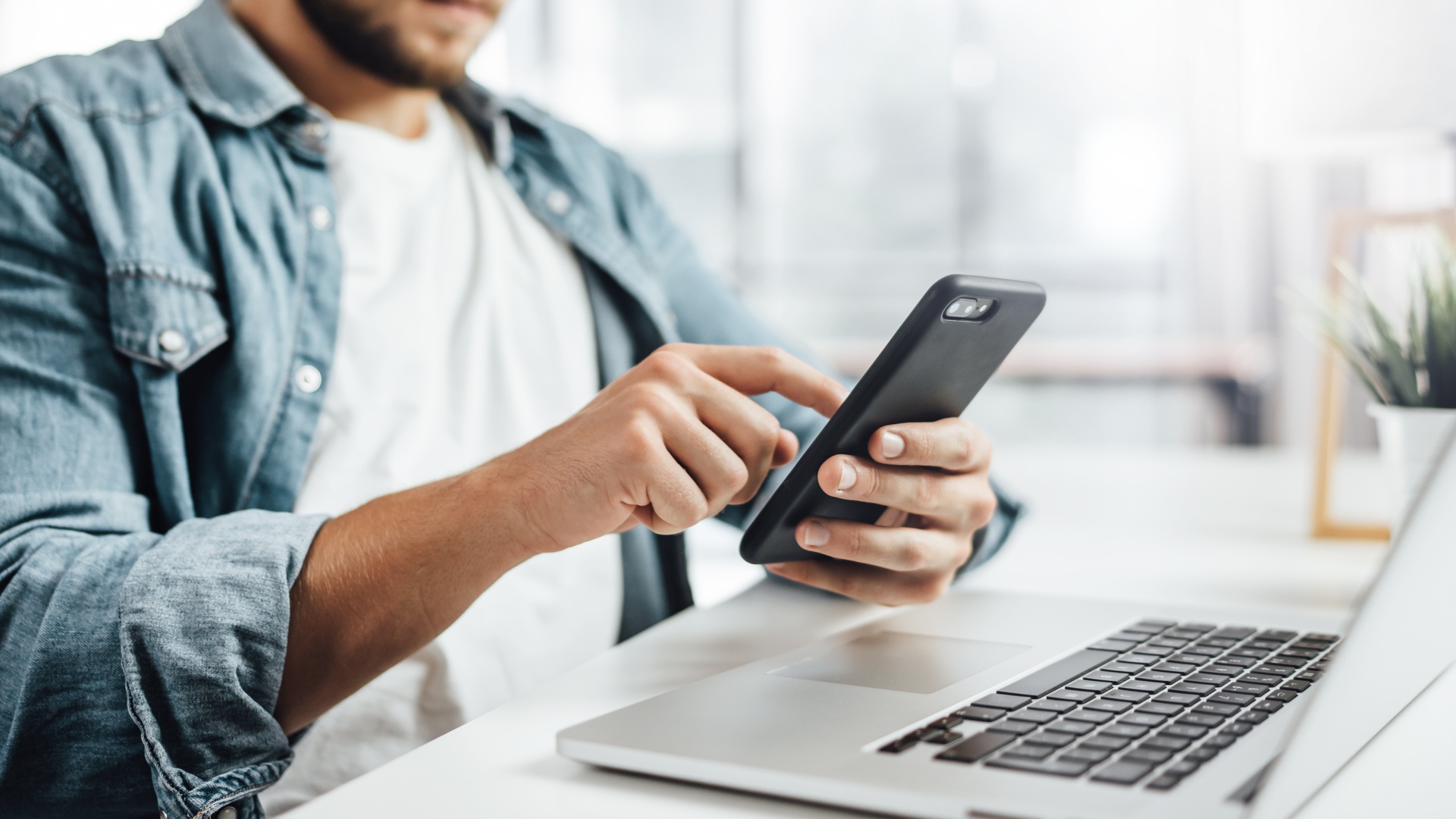 A person sits at a desk holding a black smartphone and scrolling. An open laptop sits in front of them.