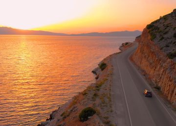 A small car driving down the road next to a large lake. The sun sets in the orange sky in the distance.