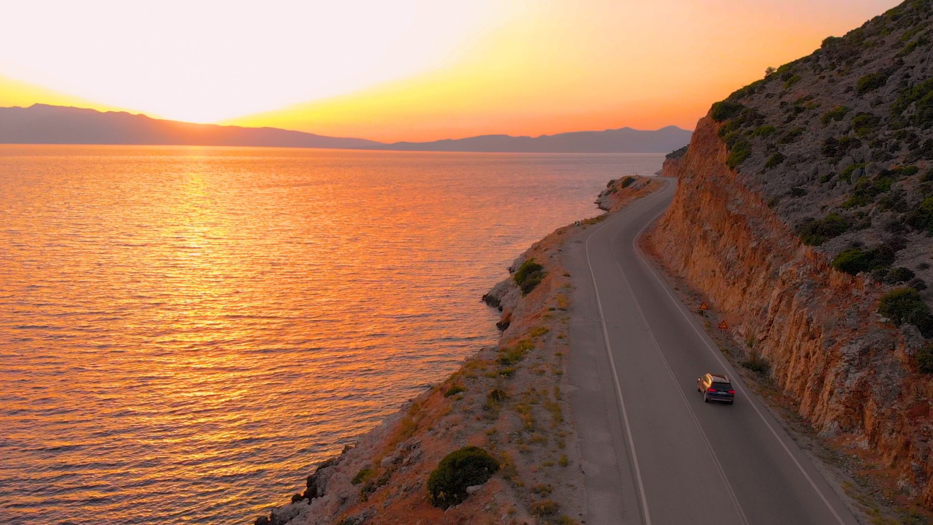 A small car driving down the road next to a large lake. The sun sets in the orange sky in the distance.