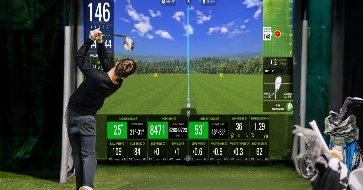 A golfer finishing a swing on indoor turf before a large golf simulator screen with data panels and equipment.