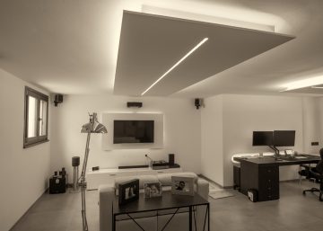 An open room features two windows, a desk with a PC and a gaming chair, a television on the wall, and a couch.