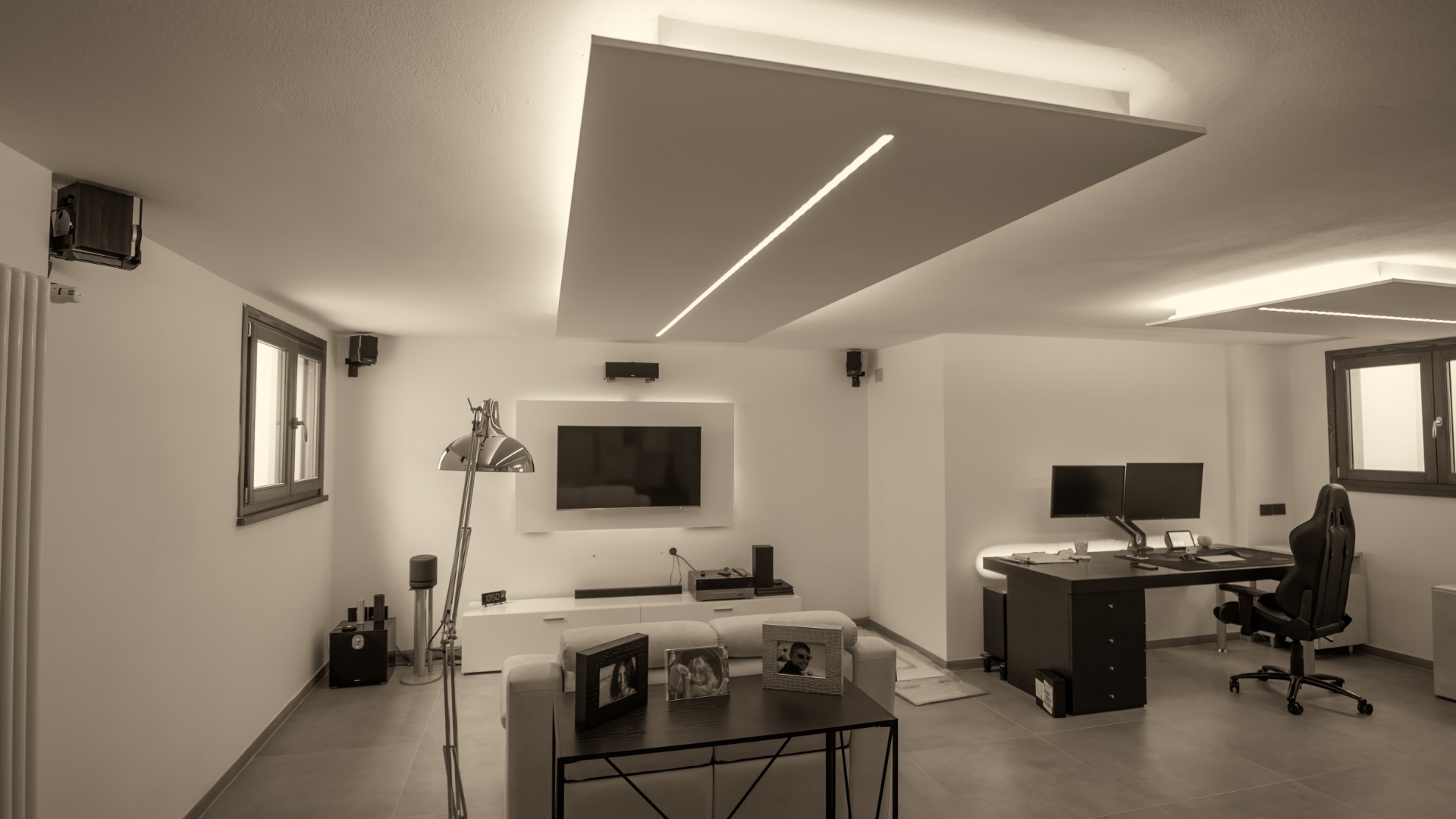 An open room features two windows, a desk with a PC and a gaming chair, a television on the wall, and a couch.