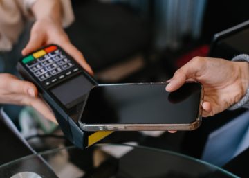 A customer holds a smartphone over a payment terminal while a cashier holds the device at a checkout counter.