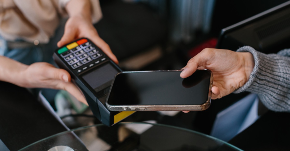 A customer holds a smartphone over a payment terminal while a cashier holds the device at a checkout counter.