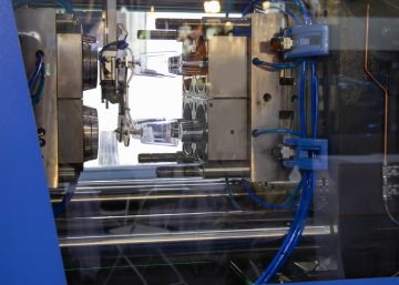 A large metal injection molding machine in a bright blue enclosure produces small, translucent plastic cups.