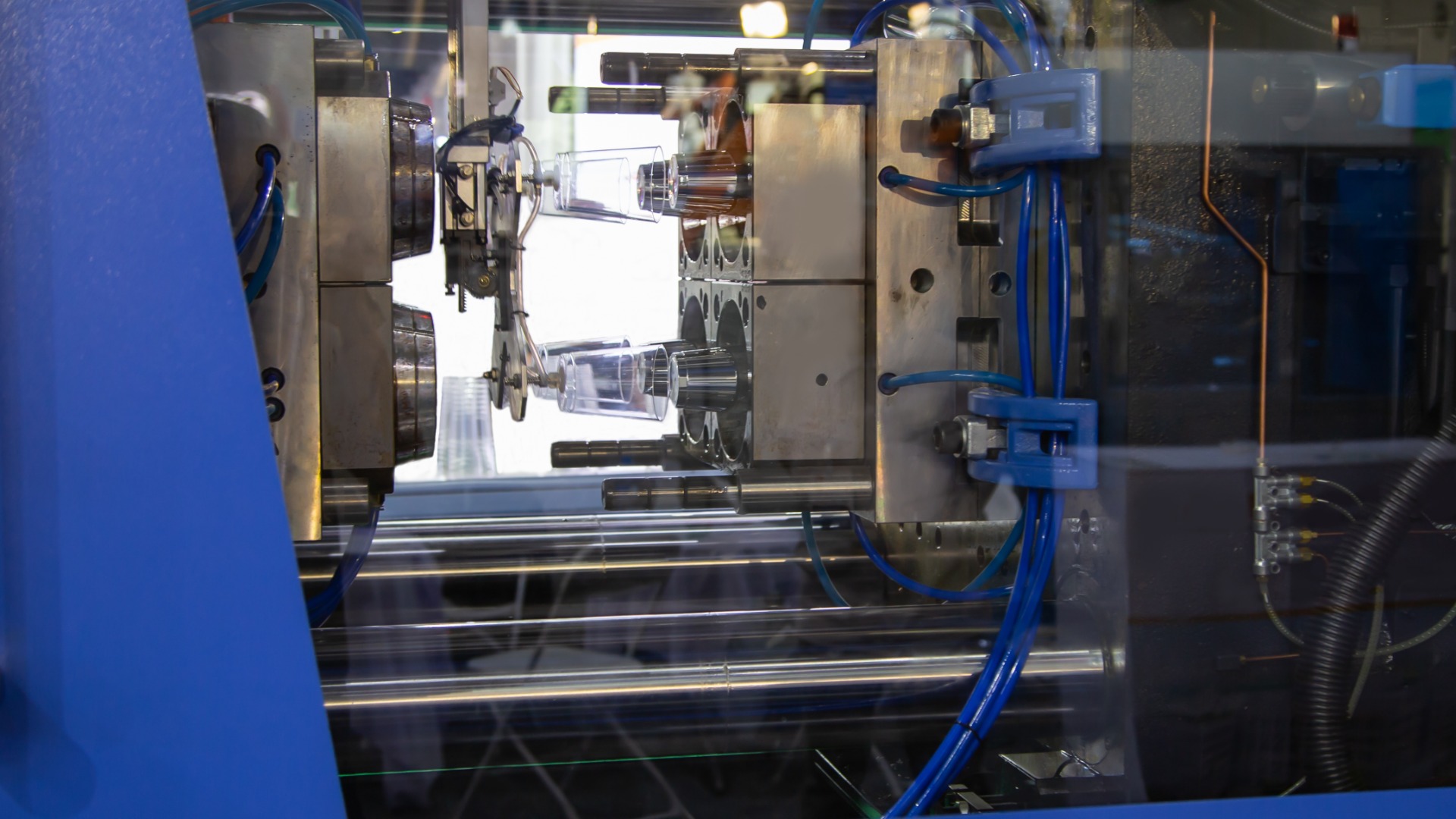 A large metal injection molding machine in a bright blue enclosure produces small, translucent plastic cups.